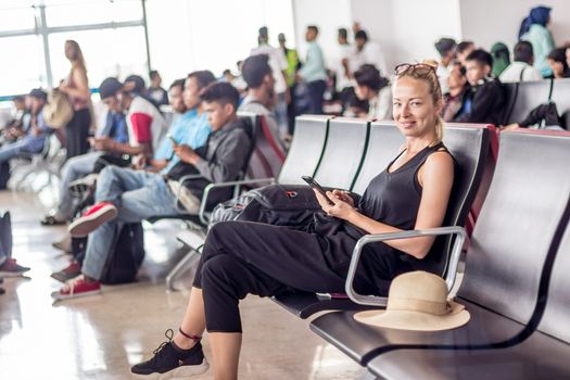 Casual sporty young blond female traveler using her cell phone while waiting to board a plane at the departure gates at the asian airport terminal.
