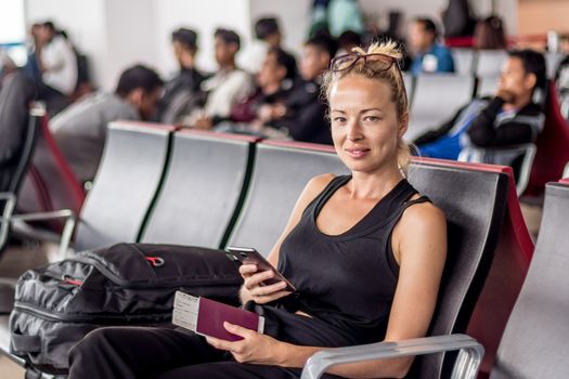 Casual tanned blond female traveler holding cell phone, passport and boarding pass while waiting to board a plane at the departure gates at the asian airport terminal.