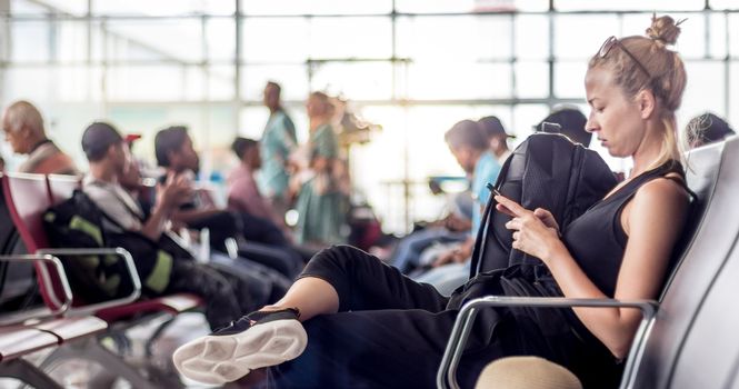 Casual sporty young blond female traveler using her cell phone while waiting to board a plane at the departure gates at the asian airport terminal.