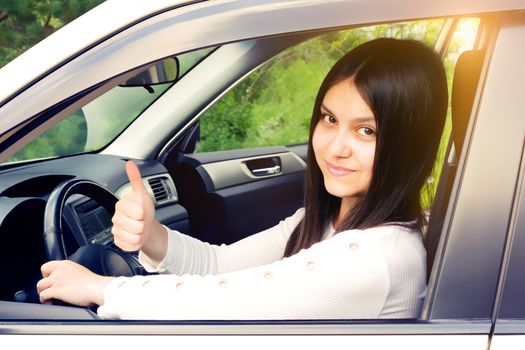 Happy young girl with long dark hair driving a car and gesturing thumb up.