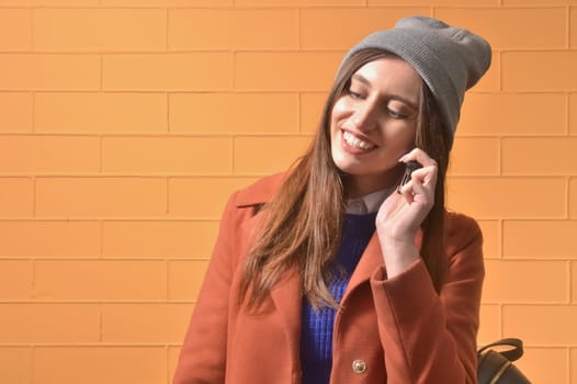 Stylish brunette in a coat and a gray cap talking on the phone on a yellow background. Young woman using a mobile phone hipster