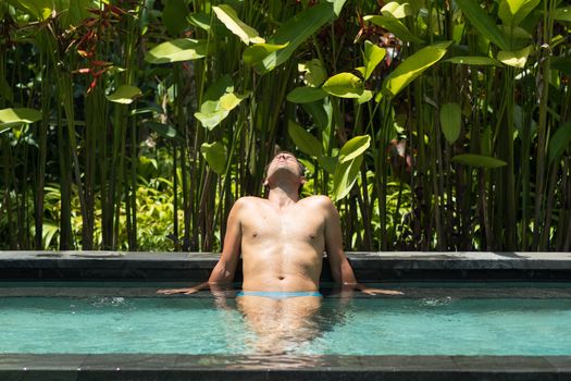 Man relaxing in outdoor spa infinity swimming pool surrounded with lush tropical greenery of Ubud, Bali. Male wellness, natural beauty and body care concept.