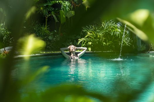 Sensual young woman relaxing in outdoor spa infinity swimming pool surrounded with lush tropical greenery of Ubud, Bali. Wellness, natural beauty and body care concept.