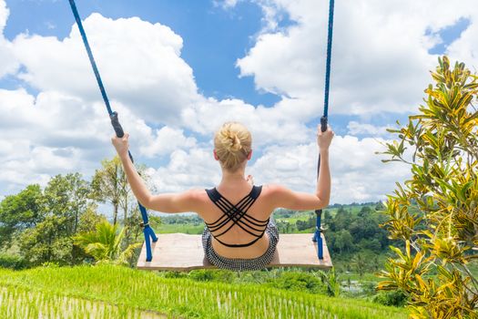 Happy sporty female traveller swinging on wooden swing, enjoying summer vacation in pristine green nature at Jatiluwih rice terraces on Bali, Indonesia. Freedom and life enjoying concept.