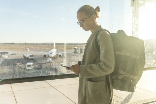 Casually dressed female traveler at airport looking at smart phone device in front of airport gate windows overlooking planes on airport runway.