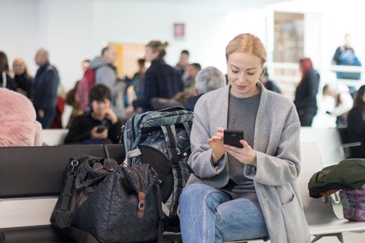 Casual blond young woman using her cell phone while waiting to board a plane at the departure gates at the airport terminal.