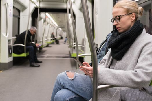 Beautiful blonde caucasian woman wearing winter coat and scarf reading on the phone while traveling by metro. Public transport.