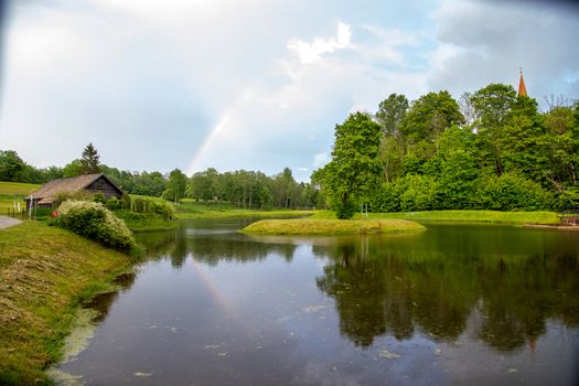 Rainbow over the pond, trees and wooden house on summer day. Rainbow and trees reflection in water, Latvia. 