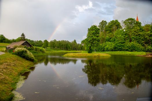 Rainbow over the pond, trees and wooden house on summer day. Rainbow and trees reflection in water, Latvia. 