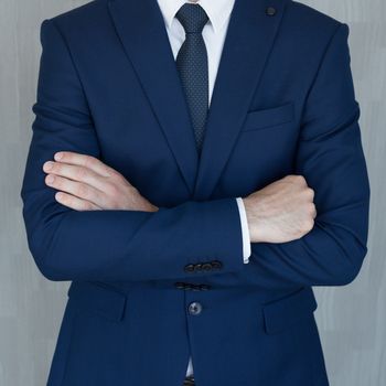 Torso of anonymous businessman standing with hands in lowered steeple wearing beautiful fashionable classic grey suit, white shirt and blue tie, against grey backgound.