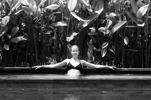 Sensual young woman relaxing in outdoor spa infinity swimming pool surrounded with lush tropical greenery of Ubud, Bali. Wellness, natural beauty and body care concept. Black and white image.