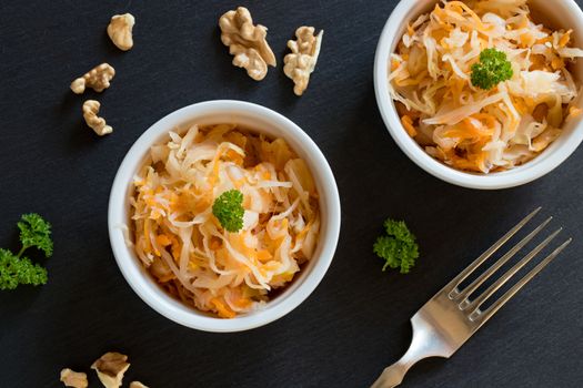 Top view of fermented cabbage and carrots in two round bowls on dark stone, with walnuts and parsley in the background