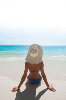 Woman in bikini and sunhat sitting on beach and looking at sea