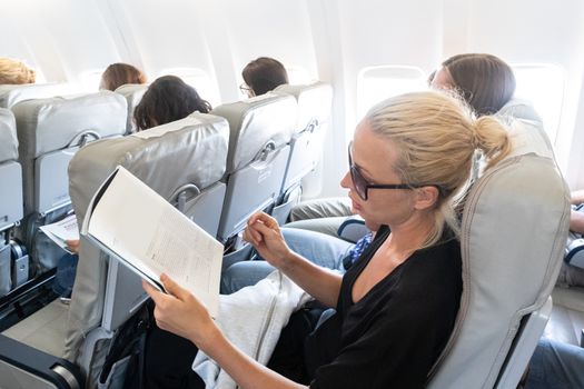 Female traveler reading magazine on airplane during flight. Female traveler reading seated in passanger cabin.