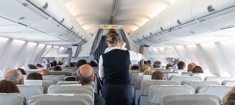 Interior of commercial airplane with flight attandant serving passengers on seats during flight. Stewardess in dark blue uniform walking the aisle. Horizontal composition.