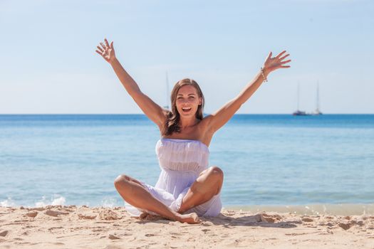 Happy woman enjoying vacation. Beautiful woman in white dress Sitting on the beach with arms outspread.