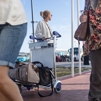 Young casual woman transporting luggage from arrival parking to international airport departure termainal by luggage trolley.