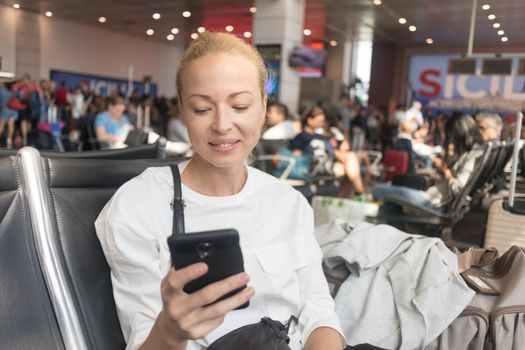 Casual blond young woman reading on her mobile phone while waiting to board a plane at the departure gates at the airport terminal. Travel concept.