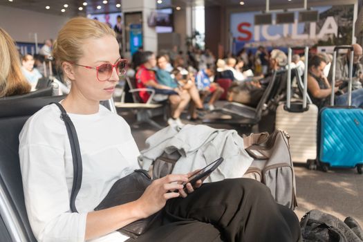 Casual blond young woman reading on her mobile phone while waiting to board a plane at the departure gates at the airport terminal. Travel concept.