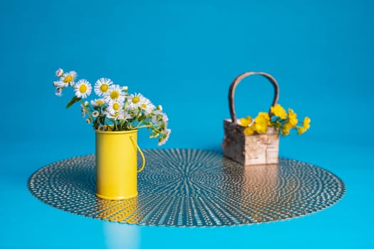 Prairie fleabane wildflowers in a small yellow cylinder vase. Hairy buttercup flowers lie in a small basket on a placemat. Teal blue blackground.