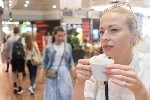 Portrait of a casual young blond woman having a cup of coffee, sitting and waiting in cafe indoors of an airport, station, food court or shopping mall.