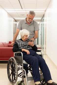 Middle aged man showing and helping elderly 95 years old woman sitting at the wheelchair how to use modern mobile phone application to monitor her therapy and entertain herself.