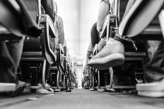 Low agle view of passenegers commercial airplane aisle with passenegers sitting on their seats while flying. Black and white image.