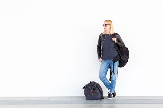 Fashionable young woman standing and waiting against plain white wall on the station whit travel bag by her side. Copy space on white wall.