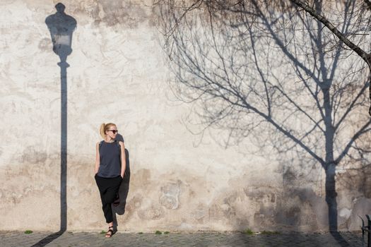 Modern trendy fashionable woman wearing sunglasses leaning against old textured retro wall. Vintage street lamp and tree shadow falling on the wall. Graphical and textured artisic image.