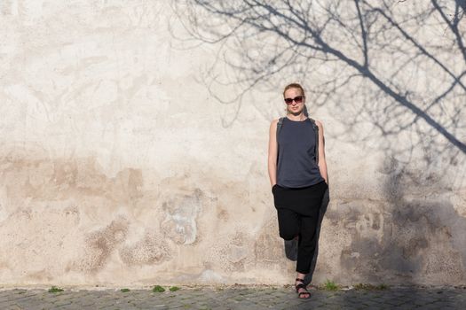 Modern trendy fashionable woman wearing sunglasses leaning against old textured retro wall with tree shadow falling on the wall. Graphical and textured artisic image.