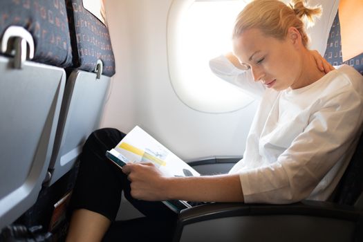 Woman feeling neck pain while reading in flight magazine on long intercontinental airplane flight. Female traveler uncomfortable seated in passenger cabin. Sun shining trough airplane window.