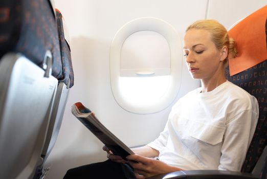 Woman reading in flight magazine on airplane. Female traveler reading seated in passanger cabin. Sun shining trough airplane window.