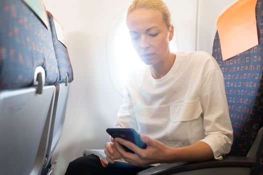 Young woman using mobile phone on airplane. Female traveler reading on her phone on the aircraft seat near the window during the flight in the airplane. Sun shining trough airplane window.