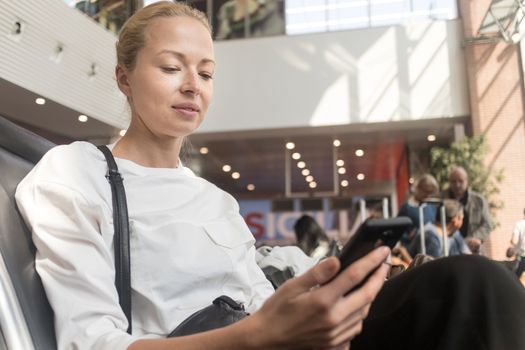 Casual blond young woman reading on her mobile phone while waiting to board a plane at the departure gates at the airport terminal. Travel concept.