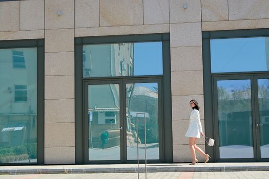 Elegant young woman dressed in a short white coat posing on a city street. Beautiful brunette woman. Modern urban woman portrait. Fashion business style clothes.