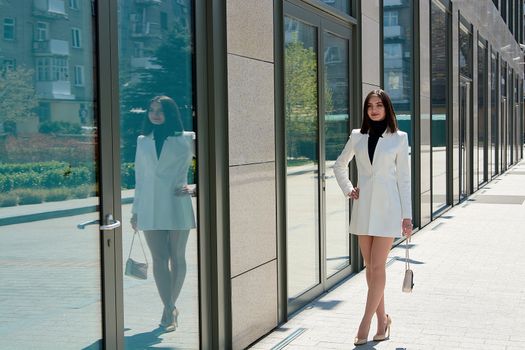 Elegant young woman dressed in a short white coat posing on a city street. Beautiful brunette woman. Modern urban woman portrait. Fashion business style clothes.