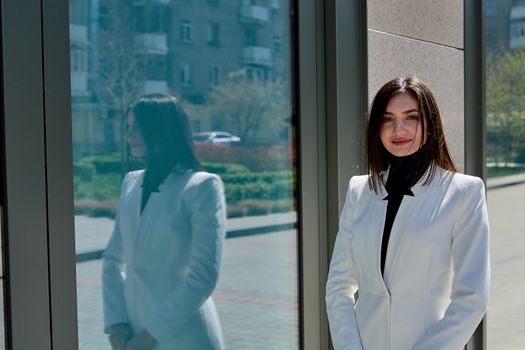 Elegant young woman dressed in a short white coat posing on a city street. Beautiful brunette woman. Modern urban woman portrait. Fashion business style clothes.