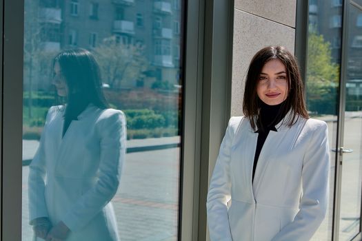 Elegant young woman dressed in a short white coat posing on a city street. Beautiful brunette woman. Modern urban woman portrait. Fashion business style clothes.