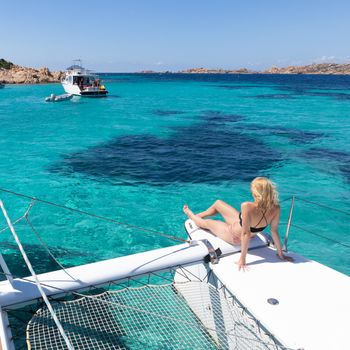 Woman in bikini tanning and relaxing on a summer sailin cruise, sitting on a luxury catamaran near picture perfect white sandy beach on Spargi island in Maddalena Archipelago, Sardinia, Italy.