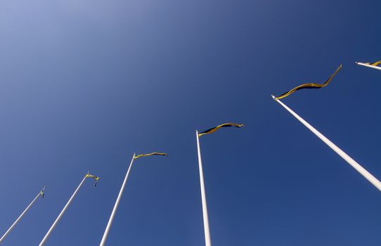 Swedish flagpoles in a diagonal row with blue and yellow pennants on a sunny day against blue sky.