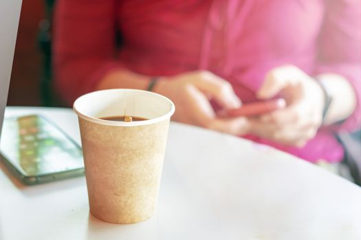 coffee in a recycled paper container with a girl checking her smart phone on the background. Blur and depth of field. Coffee break and relaxation. Wireless technology