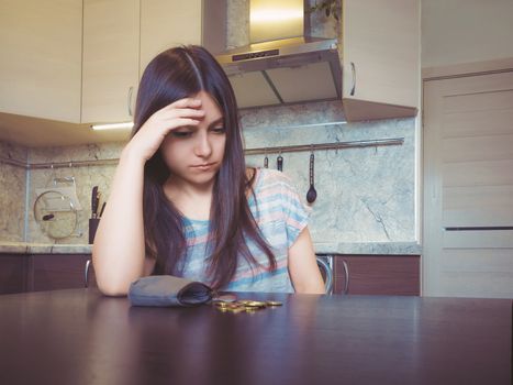 Concept of financial problems, a young disappointed woman with long dark hair, sits next to an old empty wallet with several coins on the table.