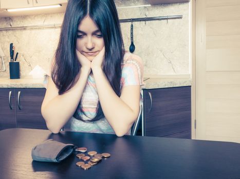 Concept of financial problems, a young disappointed woman with long dark hair, sits next to an old empty wallet with several coins on the table.