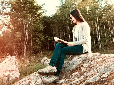 Young pretty girl reading a book sitting on a large rock in the forest.