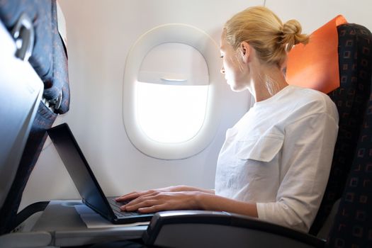 Attractive caucasian female passenger looking through the plain window while working on modern laptop computer using wireless connection on board of commercial airplane flight.