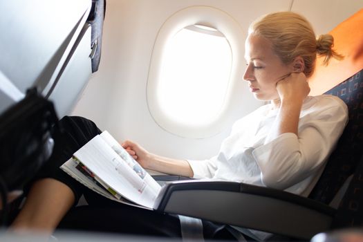 Woman reading in flight magazine on airplane. Female traveler reading seated in passanger cabin. Sun shining trough airplane window.