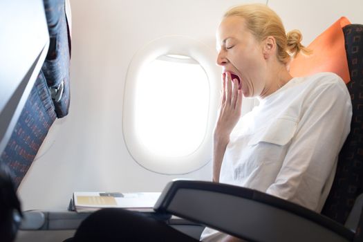 Beautiful casual caucasian woman yawning on airplane flight while reading in flight magazine. Female traveler reading seated in passanger cabin. Sun shining trough airplane window.