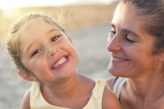 portrait of a girl smiling with her mother, the girl looks at camera while her mother smile looking at her daughter