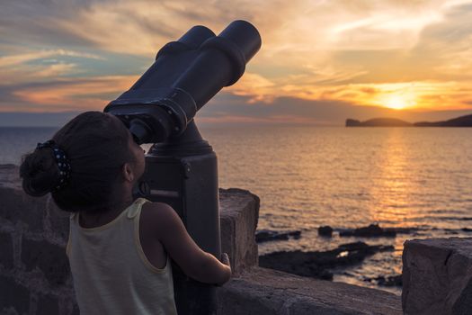 little girl looking through a binoculars for tourists during the sunset in front of the giant that sleeps in the Gulf of Alghero, Sardinia, Italy