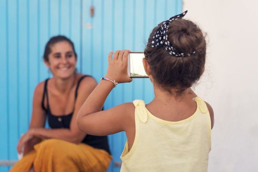 little girl taking pictures with the smart phone to her mother in front of a blue door, single-parent family on vacation, divorced woman enjoying with her daughter